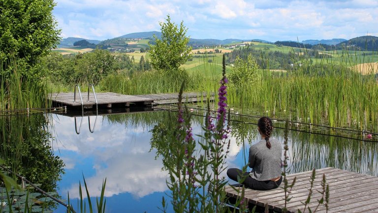 Frau sitzt auf kleinem Steg am Teich und schaut in die Hügellandschaft 