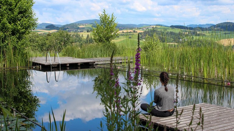 Frau sitzt auf kleinem Steg am Teich und schaut in die Hügellandschaft